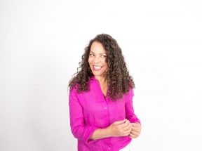 Eine Frau mit langen lockigen Haaren und einer leuchtend rosa Bluse lächelt und blickt vor einem schlichten weißen Hintergrund zur Seite.