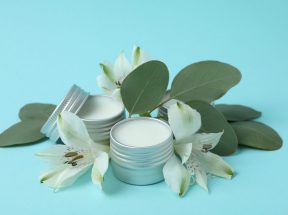 Kleine offene Metalldosen, gefüllt mit weißer Creme, stehen zwischen weißen Blumen und grünen Blättern auf hellblauem Hintergrund und sorgen für ein frisches und natürliches Aussehen.