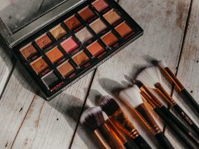 Eine offene Lidschattenpalette mit 18 warmen Farbtönen liegt neben sieben Make-up-Pinseln auf einer Holzoberfläche. Denken Sie daran, den Pinsel regelmäßig zu reinigen, um eine gesündere Haut und ein makelloses Auftragen zu gewährleisten.