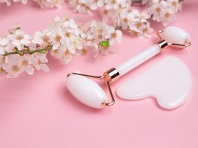 Ein Gesichtsroller aus weißer Jade und ein Gua Sha-Gesichtsmassagestein liegen auf einer rosa Oberfläche neben blühenden weißen Kirschblüten und schaffen eine heitere Szene, die Schönheit und Selbstfürsorge hervorruft.