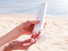 Eine Person drückt Sonnenschutz Make-up aus einer weißen Tube auf ihre Hand an einem Sandstrand, mit dem Meer und dem blauen Himmel im Hintergrund.